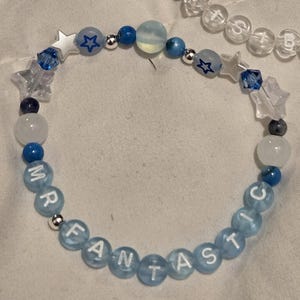 Op de afbeelding: Twee handgemaakte armbanden met kralen. E&eacute;n armband heeft doorzichtige kralen met de tekst "BELIEVE" en blauwe ster- en parelaccenten. De andere armband heeft blauwe kralen met de tekst "MR FANTASTIC" met ster- en ronde kralenaccenten. Beide armbanden liggen op een wit oppervlak.