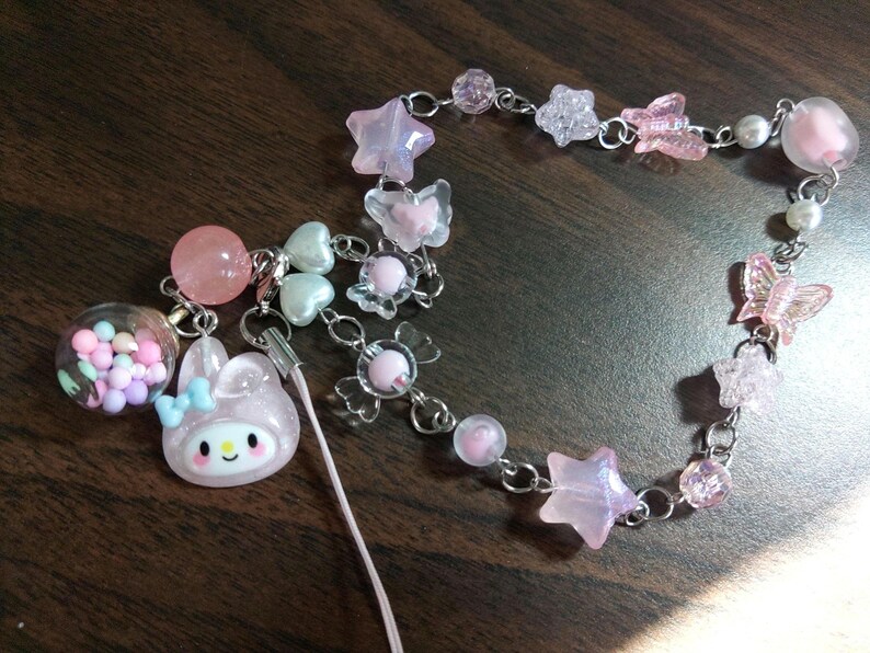 May include: A pink and white charm bracelet with a variety of charms, including stars, butterflies, and a small pink bunny. The bracelet is made of silver chain and has a pink and white string attached to it.