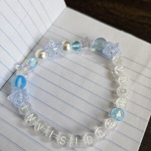 Op de afbeelding: Een doorzichtige kralenarmband met het woord "INVISIBLE" in witte letterkralen. De armband heeft blauwe en doorzichtige kralen, waaronder ster- en ronde vormen. Een hartvormige kraal is ook zichtbaar.