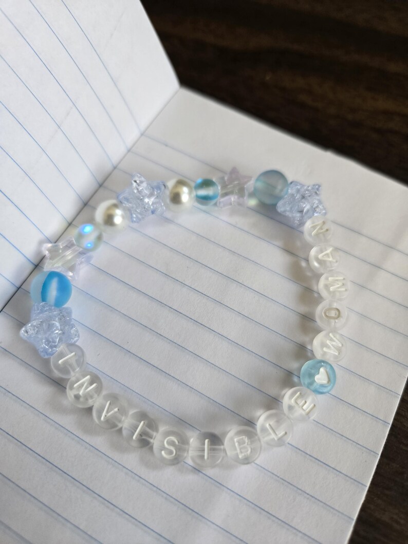 Op de afbeelding: Een doorzichtige kralenarmband met het woord "INVISIBLE" in witte letterkralen. De armband heeft blauwe en doorzichtige kralen, waaronder ster- en ronde vormen. Een hartvormige kraal is ook zichtbaar.