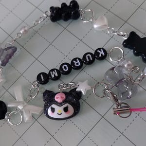 May include: A black and white charm bracelet with a black and white Hello Kitty charm, black bear charms, and a silver charm with the word "KUROMI" spelled out.