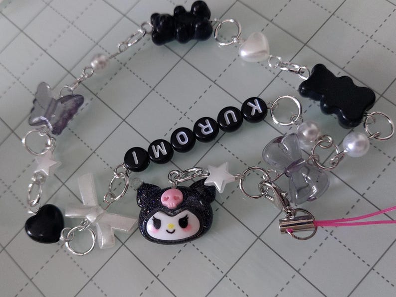 May include: A black and white charm bracelet with a black and white Hello Kitty charm, black bear charms, and a silver charm with the word "KUROMI" spelled out.