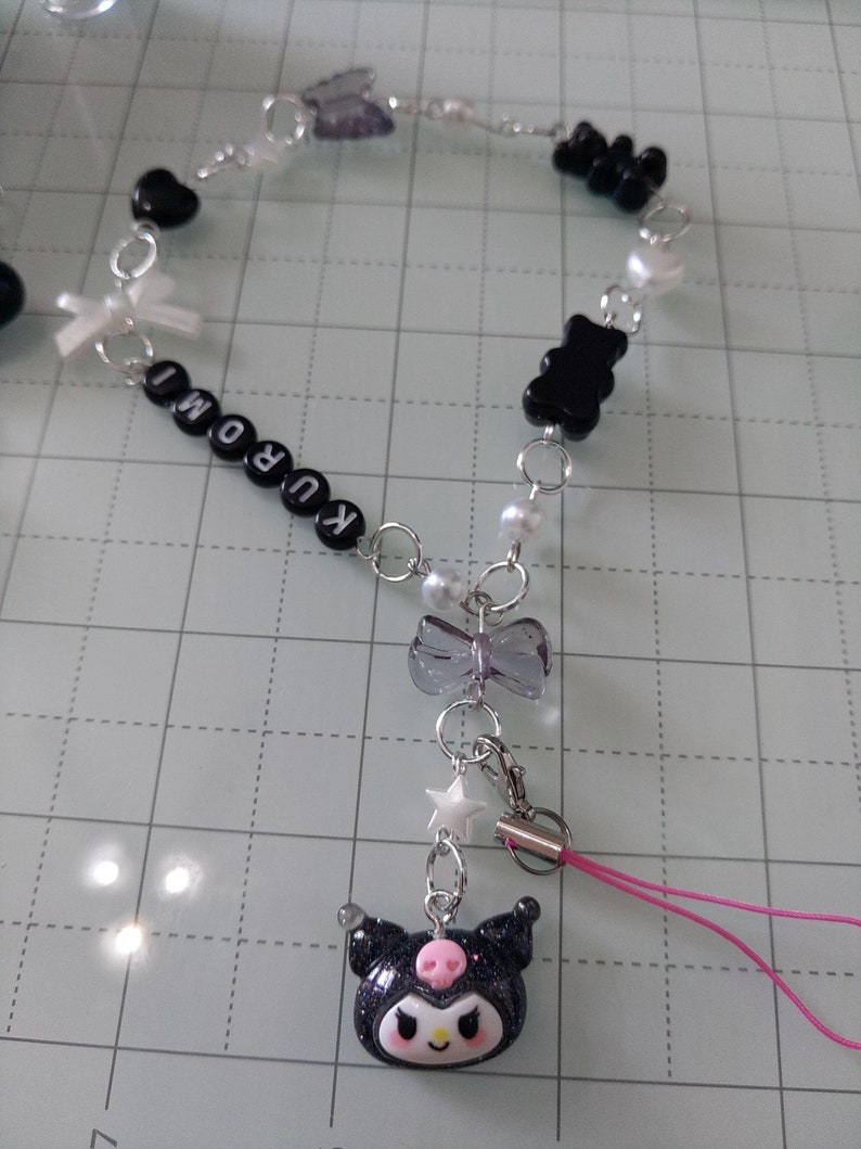 May include: Black and white beaded charm bracelet with a black and white Kuromi charm attached to a pink phone strap. The bracelet features black bears, hearts, bows, and white pearls.