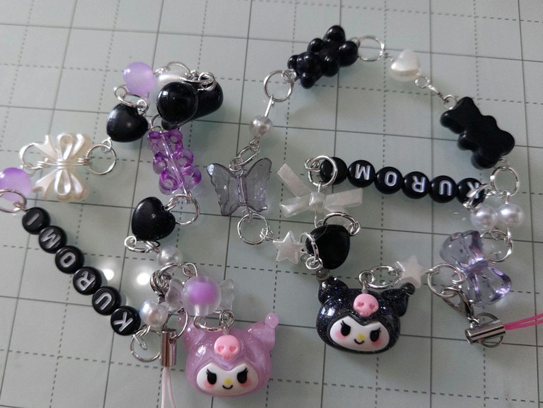 May include: A black and white phone charm with a black and white Hello Kitty character, a purple and white butterfly, and a black and white heart. The charm also includes black and white beads that spell out "KUROMI".