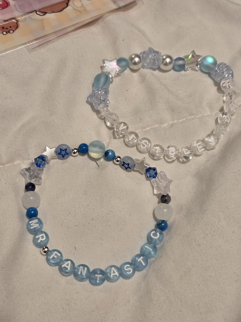 Op de afbeelding: Twee handgemaakte armbanden met kralen. E&eacute;n armband heeft doorzichtige kralen met de tekst "BELIEVE" en blauwe ster- en parelaccenten. De andere armband heeft blauwe kralen met de tekst "MR FANTASTIC" met ster- en ronde kralenaccenten. Beide armbanden liggen op een wit oppervlak.