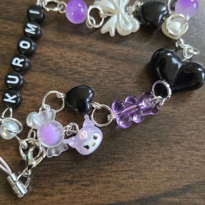 May include: A purple, black, and white charm bracelet with the word "KUROMI" spelled out in black beads. The bracelet features a variety of charms, including hearts, bows, and a gummy bear.