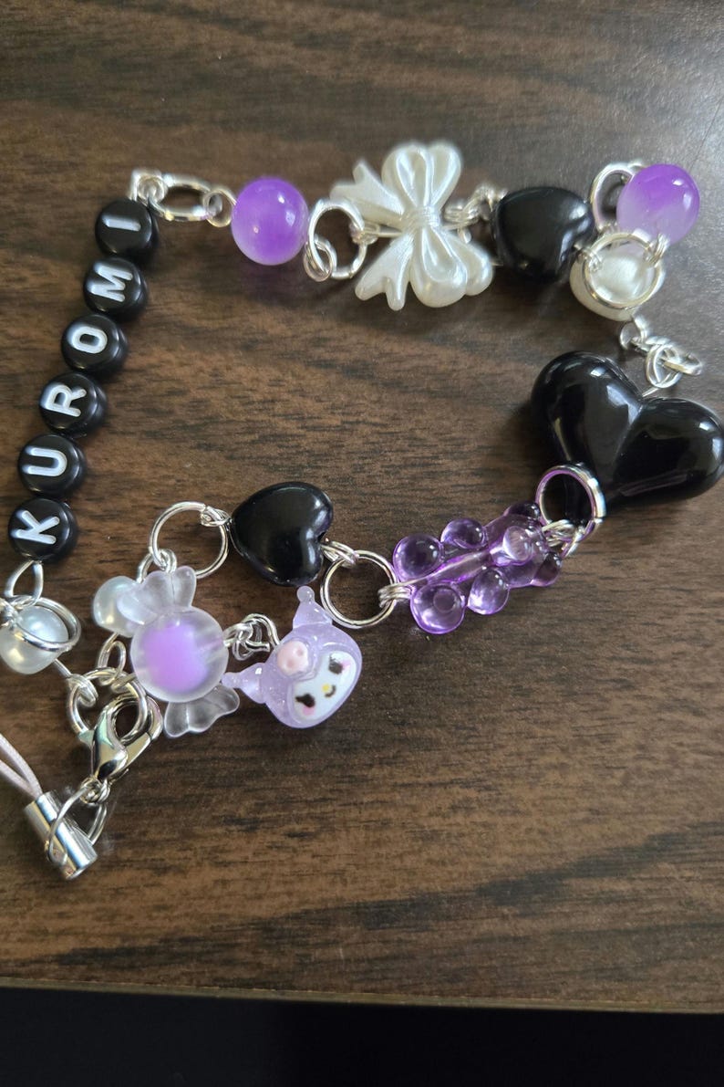 May include: A purple, black, and white charm bracelet with the word "KUROMI" spelled out in black beads. The bracelet features a variety of charms, including hearts, bows, and a gummy bear.