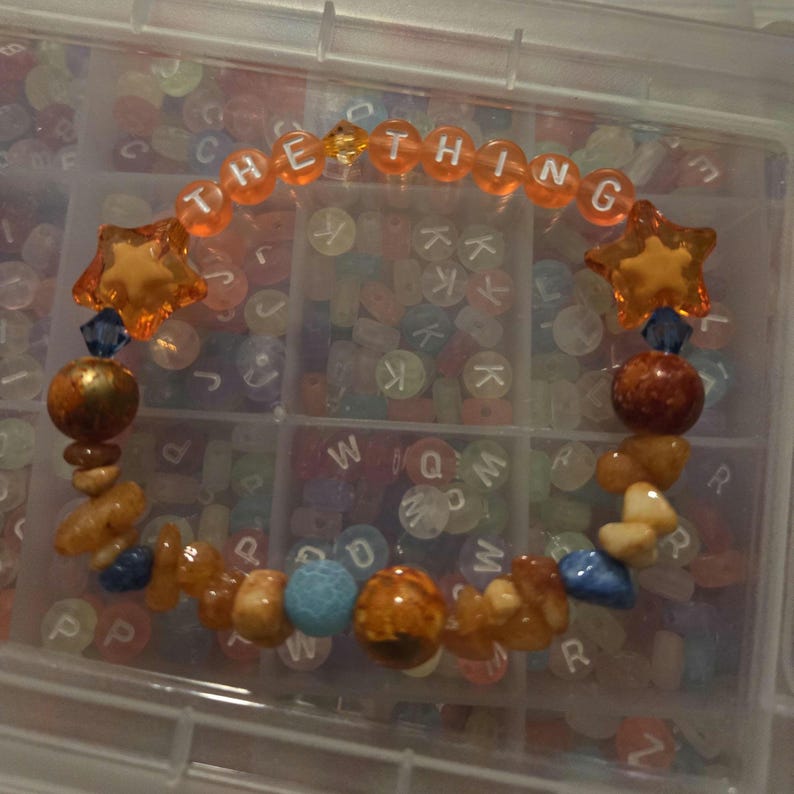 Op de afbeelding: Een handgemaakte armband met de tekst "THE THING" gespeld in oranje letterkralen. De armband heeft oranje stervormige kralen, blauwe en oranje kralen en diverse andere kleurrijke stenen. De armband wordt gepresenteerd in een doorzichtige plastic container.