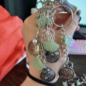May include: A collection of silver keychains with zodiac sign charms. Each keychain features a large ring, a lobster clasp, and decorative charms, including green seashell-shaped beads and zodiac symbols like Aries and Taurus.