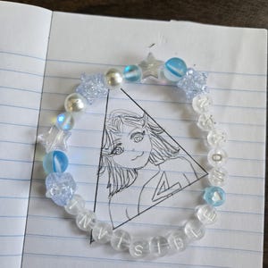 Op de afbeelding: Een kralenarmband met doorzichtige en blauwe kralen, waaronder stervormige en ronde kralen. De armband spelt "INVISIBLE" en "NOW" met letterkralen. De armband ligt op een notitieboekpagina met een tekening.