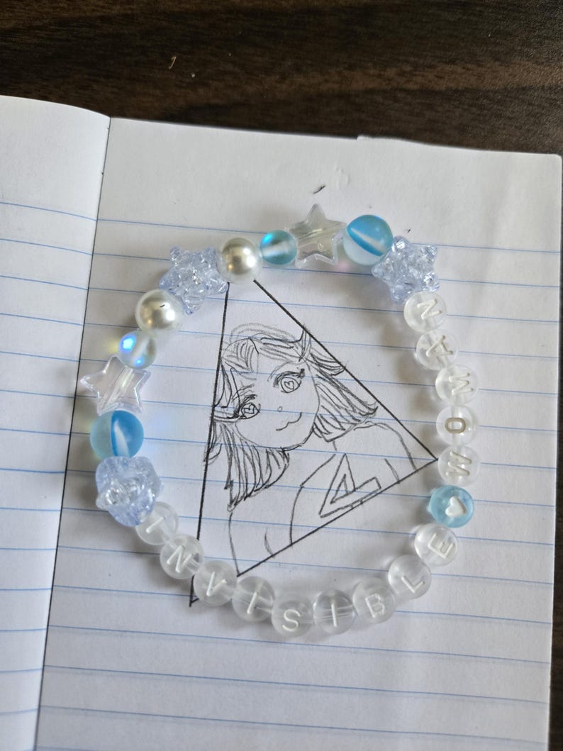 Op de afbeelding: Een kralenarmband met doorzichtige en blauwe kralen, waaronder stervormige en ronde kralen. De armband spelt "INVISIBLE" en "NOW" met letterkralen. De armband ligt op een notitieboekpagina met een tekening.