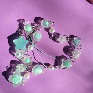 Puede incluir: Un brazalete de charms azul claro y transparente con una variedad de charms, incluyendo estrellas, flores, corazones y un caramelo. El brazalete está unido a una funda de teléfono.
