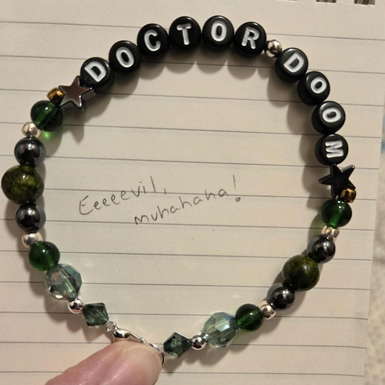 Op de afbeelding: Een kralenarmband met de woorden "DOCTOR DOOM" in zwarte letterkralen. De armband heeft groene, zilveren en zwarte kralen, met stervormige accenten. De handgeschreven tekst "Eeeevil, muhahaha!" is ook zichtbaar.