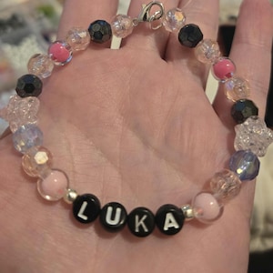 Op de afbeelding: Een kralenarmband met de naam "LUKA" gespeld in zwarte en witte letterkralen. De armband heeft een verscheidenheid aan kralen in roze, heldere, zwarte en iriserende kleuren. Een zilveren sluiting zet de armband vast.