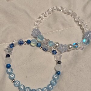 Op de afbeelding: Twee armbanden met kralen. E&eacute;n armband heeft de tekst "MR FANTASTIC" in lichtblauwe kralen. De andere armband heeft de tekst "SISTER SOUL" in doorzichtige kralen. Beide armbanden hebben blauwe en witte kralen, stervormige kralen en parels.