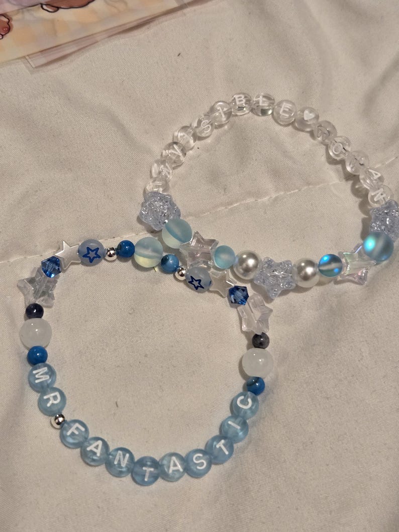 Op de afbeelding: Twee armbanden met kralen. E&eacute;n armband heeft de tekst "MR FANTASTIC" in lichtblauwe kralen. De andere armband heeft de tekst "SISTER SOUL" in doorzichtige kralen. Beide armbanden hebben blauwe en witte kralen, stervormige kralen en parels.