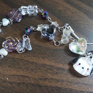 May include: A purple and white charm bracelet with a clear heart charm, a flower charm, and a small dog charm. The bracelet is attached to a clear phone strap with a white heart charm.