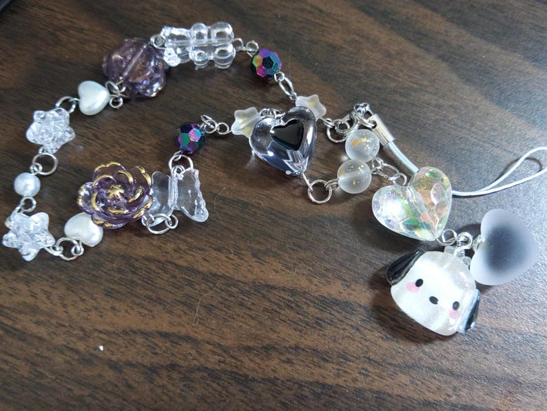 May include: A purple and white charm bracelet with a clear heart charm, a flower charm, and a small dog charm. The bracelet is attached to a clear phone strap with a white heart charm.