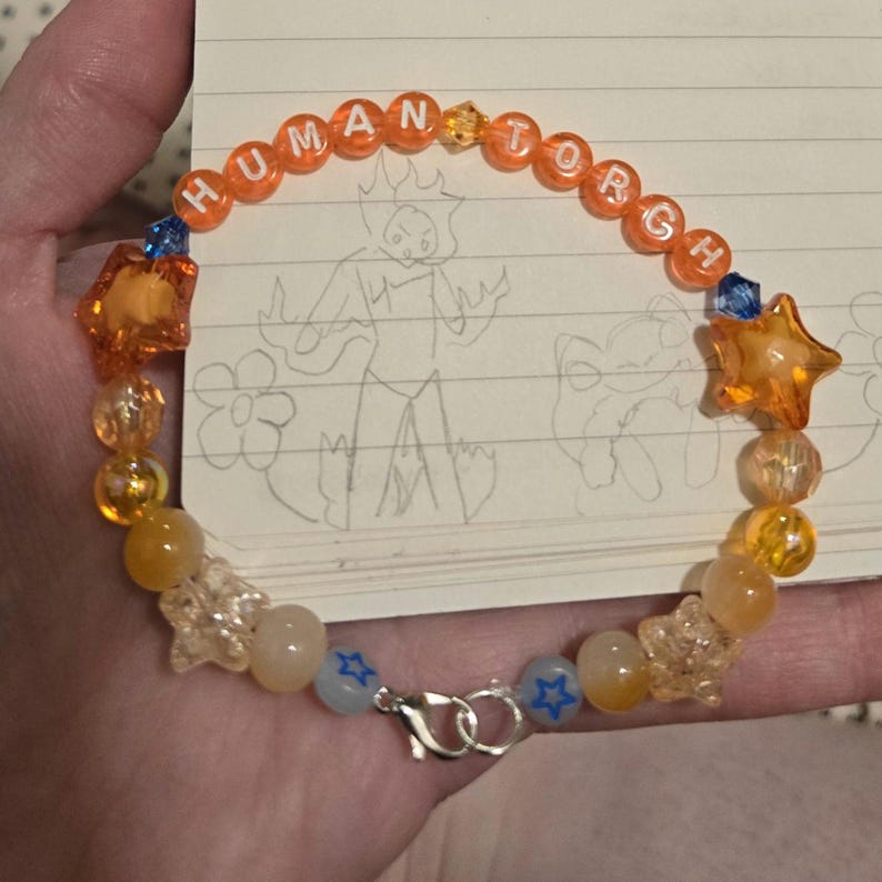 Op de afbeelding: Een kralenarmband met het woord "HUMAN TORCH" gespeld in oranje letterkralen. De armband heeft oranje stervormige kralen, heldere stervormige kralen en andere oranje en gele kralen. Een zilveren sluiting zet de armband vast.