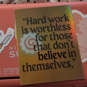 May include: A gold sticker with the quote "Hard work is worthless for those that don't believe in themselves." The sticker is on a pink surface with other stickers, including a knife and a cartoon axolotl.