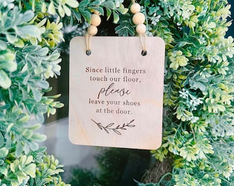 May include: Wooden sign with the text "Since little fingers touch our floor, please leave your shoes at the door." hanging on a green leafy wreath.
