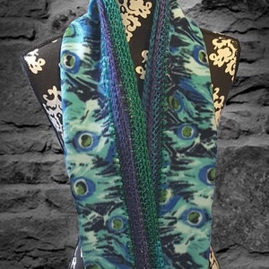 May include: A peacock-themed infinity scarf displayed on a mannequin. The scarf features a teal and blue pattern with green and purple accents. The edges are trimmed with a knitted border. The mannequin is black with white floral designs.