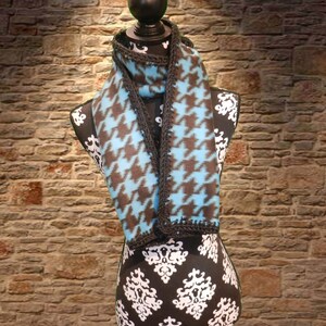 May include: A blue and brown patterned fleece scarf with a black crocheted trim. The scarf is displayed on a black and white patterned mannequin.