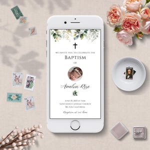 Puede incluir: Un smartphone blanco muestra una invitación de bautizo con el texto "WE INVITE YOU TO CELEBRATE THE BAPTISM OF Amelia Rose." La invitación incluye una foto de bebé y detalles florales. El teléfono está rodeado de sellos, flores y un cuenco pequeño.