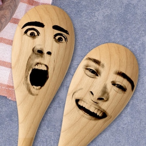 Może przedstawiać: Dwie drewniane łyżki kuchenne z wygrawerowanymi twarzami. Jedna łyżka przedstawia zaskoczoną minę z szeroko otwartymi oczami i ustami. Druga łyżka pokazuje uśmiechniętą twarz. Łyżki są wykonane z jasnego drewna.
