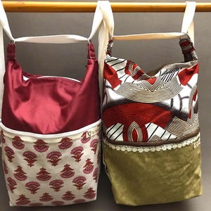 Könnte beinhalten: Zwei Stofftaschen hängen an einer Holzstange. Die Tasche links ist rot mit einem weißen und roten Blumendruck am Boden. Die Tasche rechts hat einen weißen, roten und braunen geometrischen Druck mit einer braunen und weißen Spitzenborte und einem grünen Boden.