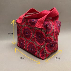 Pode incluir: Uma bolsa tote de tecido vermelho com um padrão circular repetido em roxo e dourado. A bolsa tem fecho de correr e alças vermelhas. A bolsa mede aproximadamente 17 cm de comprimento, 14 cm de altura e 10 cm de largura.