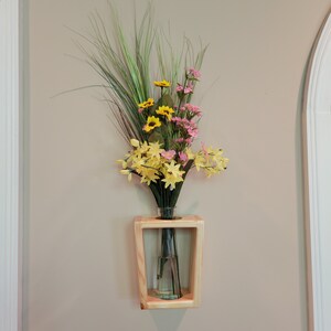 May include: A wooden wall-mounted vase with a clear glass bottle holding an arrangement of colourful artificial flowers. The flowers include yellow, pink, and green elements, creating a vibrant display. The vase is made of light-coloured wood.