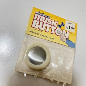 May include: A vintage electronic music button in a clear plastic package. The button is round with a reflective center and a cream-colored border. The packaging is yellow and features the text "electronic MUSIC BUTTON" and "Wedding March".