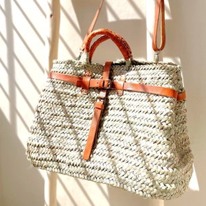 May include: A large woven straw tote bag with a brown leather strap and buckle. The bag has a woven design and is hanging on a wooden ladder.