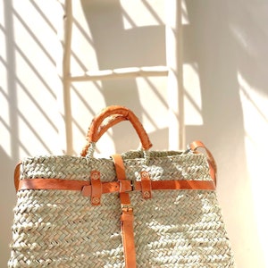 May include: A large, woven straw tote bag with brown leather straps and buckles. The bag has a wide, rectangular shape and is perfect for carrying everyday essentials.