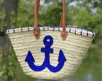May include: A white straw tote bag with blue trim and a large blue anchor design. The bag has brown leather handles.