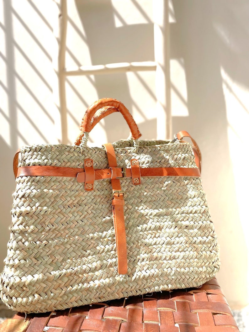 May include: A large, woven straw tote bag with brown leather straps and buckles. The bag has a wide, rectangular shape and is perfect for carrying everyday essentials.
