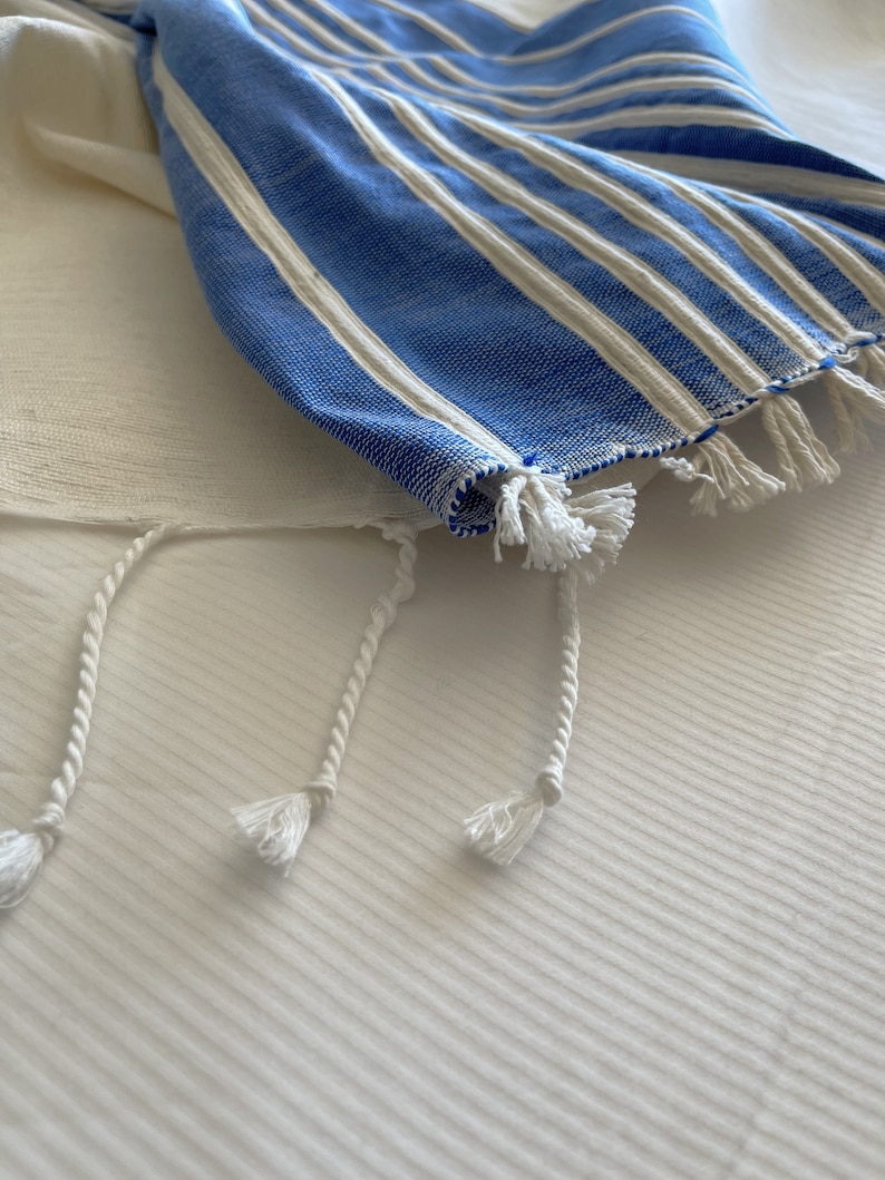 Puede incluir: Un chal de oraci&oacute;n a rayas azules y blancas con borlas blancas. El chal est&aacute; doblado y sobre una superficie blanca.