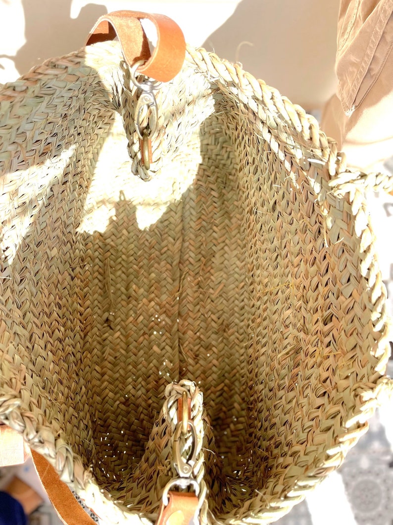 May include: A woven straw tote bag with a brown leather handle and a metal clasp. The bag is empty and the inside is visible.
