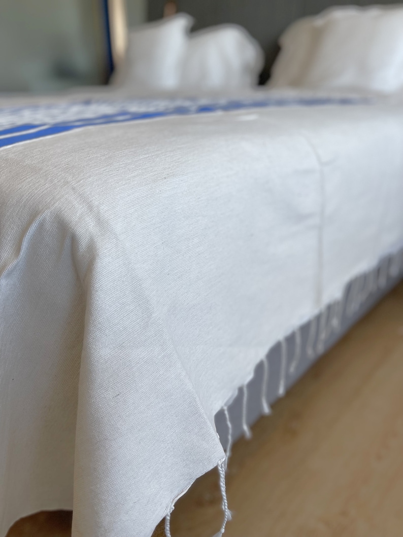 Puede incluir: Un cubrecama blanco con una franja azul y detalles de flecos. El cubrecama est&aacute; en una cama con almohadas blancas.