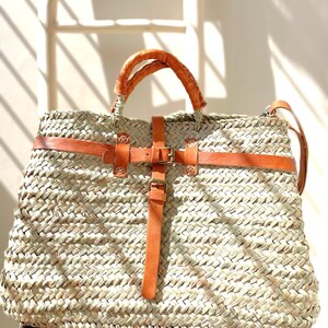 May include: A large, woven straw tote bag with brown leather handles and a buckle closure. The bag has a light brown leather strap that can be worn over the shoulder.