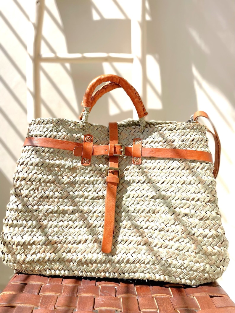 May include: A large, woven straw tote bag with brown leather handles and a buckle closure. The bag has a light brown leather strap that can be worn over the shoulder.