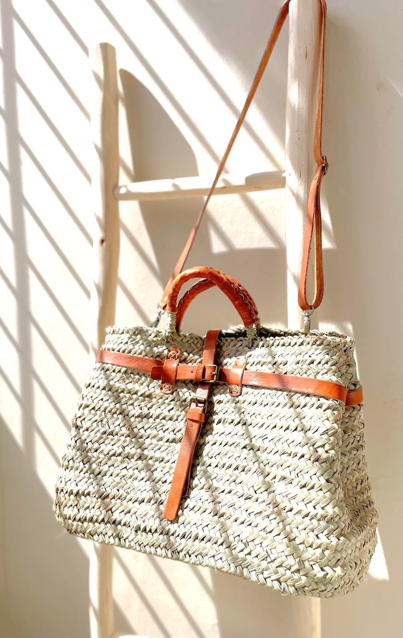 May include: A large woven straw tote bag with a brown leather strap and buckle. The bag has a woven design and is hanging on a wooden ladder.