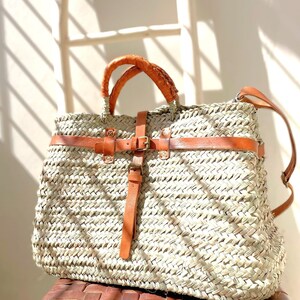 May include: A large, woven straw tote bag with brown leather straps and a buckle closure. The bag has a light brown color and is made of natural materials.