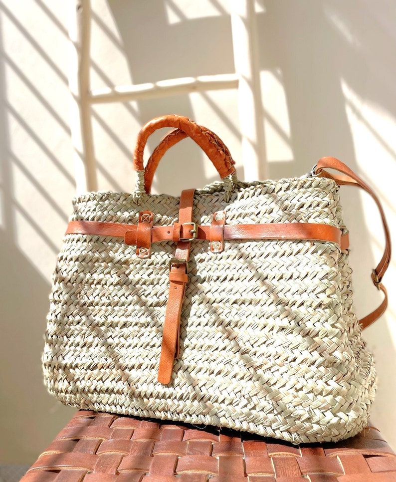 May include: A large, woven straw tote bag with brown leather straps and a buckle closure. The bag has a light brown color and is made of natural materials.