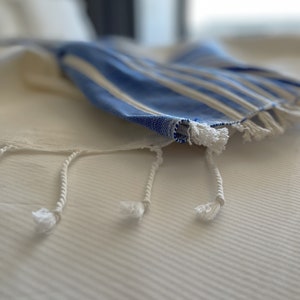 Puede incluir: Una toalla a rayas blancas y azules con flecos blancos sobre una cama blanca.