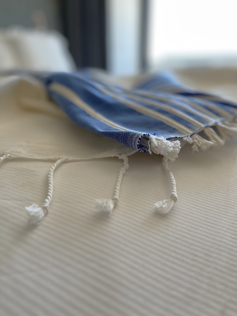 Puede incluir: Una toalla a rayas blancas y azules con flecos blancos sobre una cama blanca.