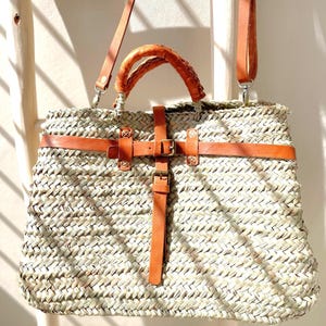 May include: A large woven straw tote bag with brown leather handles, a wide leather strap, and a buckle closure.