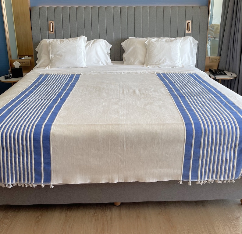 Puede incluir: Una cama blanca con una colcha a rayas azules y blancas. La cama tiene un cabecero con una cubierta de tela gris y un marco de madera.