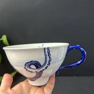 May include: White ceramic teacup with a blue octopus tentacle design. The cup has a blue handle and a blue and white painted sky design. The cup is held in a hand against a dark background.